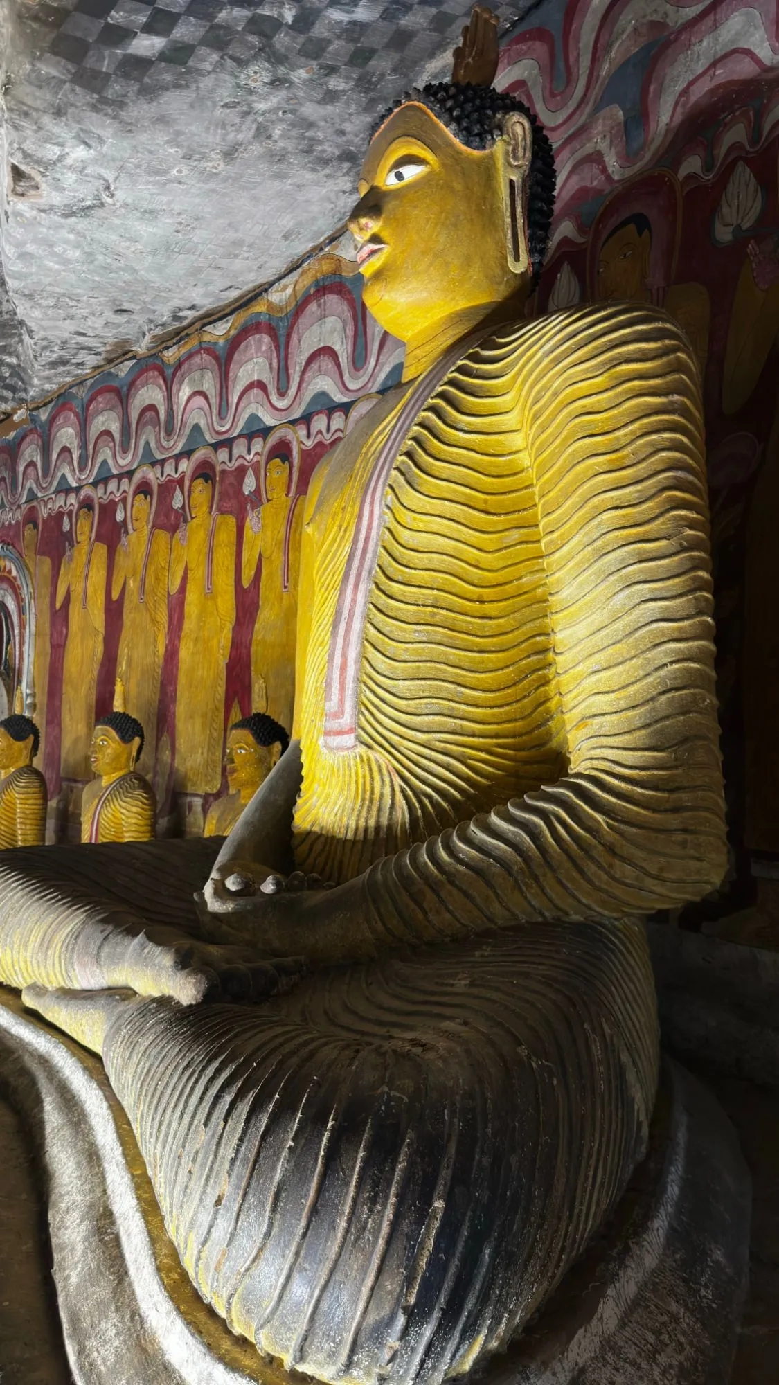 a painted golden buddha in a rock cave, under a ceiling of faded murals