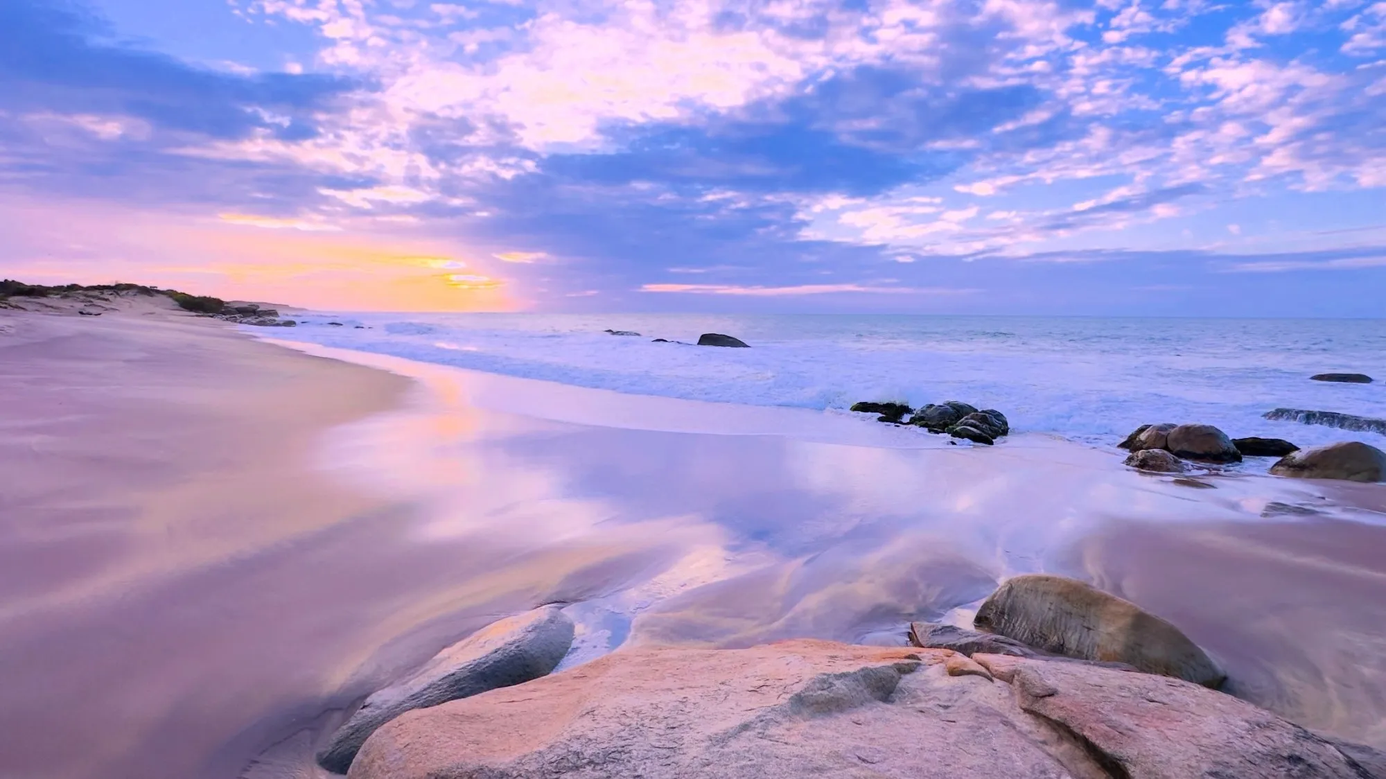 a long-exposure shore at dusk, the water smoothed to lilac over flat rocks